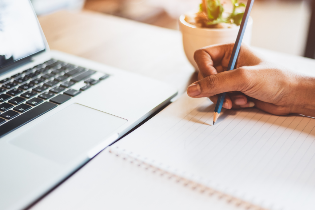 A woman is holding a pen in her hand and starts to write something in a notebook. In front of her is a laptop.