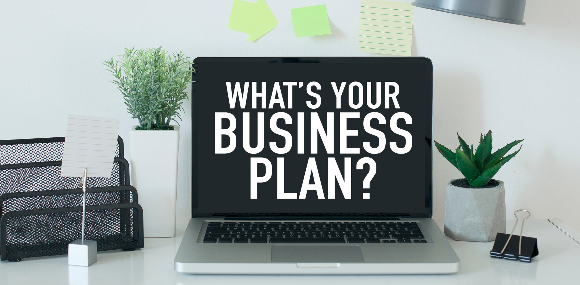 A computer is on a desk next to plants, a lamp and some pens. On the screen it is written "What's your next Business Plan?"