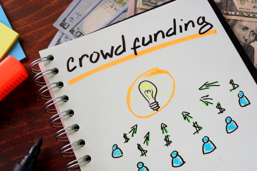 A book is placed on a desk next to some pens, sticky notes and money. On the paper you can read "crowdfunding" and underneath people with dollar signs are outlined. From there arrows point to a lightbulb.