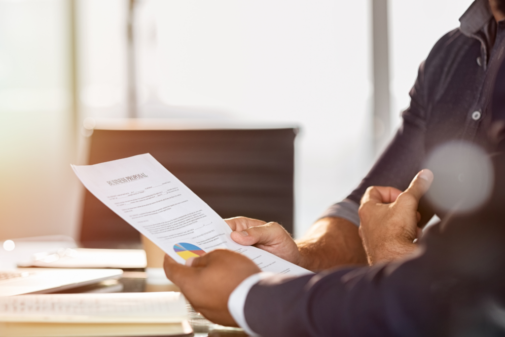 A man is holding a business proposal in his hands and sits next to another person at a table.