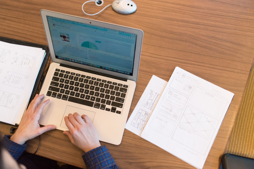 You see the hands of someone sitting at a desk. In front of him is a laptop as well as some documents.